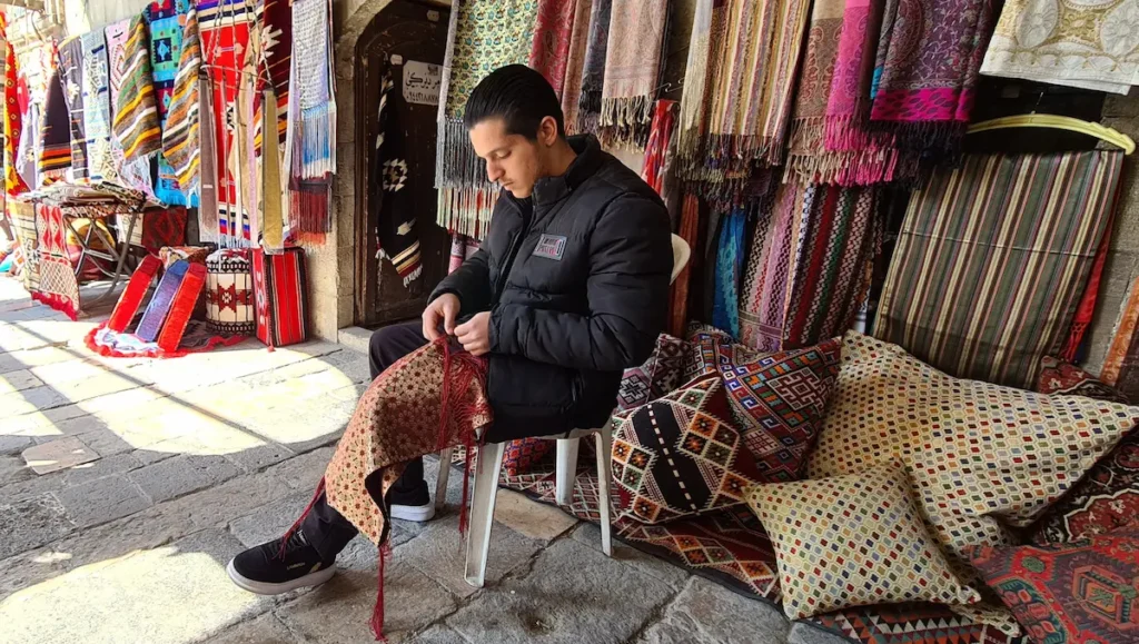 A young man sitting on a white chair in an outdoor market stall, focused on hand-stitching a patterned red and gold fabric, surrounded by colorful hanging textiles, rugs, and patterned pillows.
