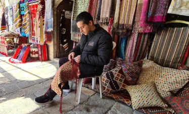 A young man sitting on a white chair in an outdoor market stall, focused on hand-stitching a patterned red and gold fabric, surrounded by colorful hanging textiles, rugs, and patterned pillows.