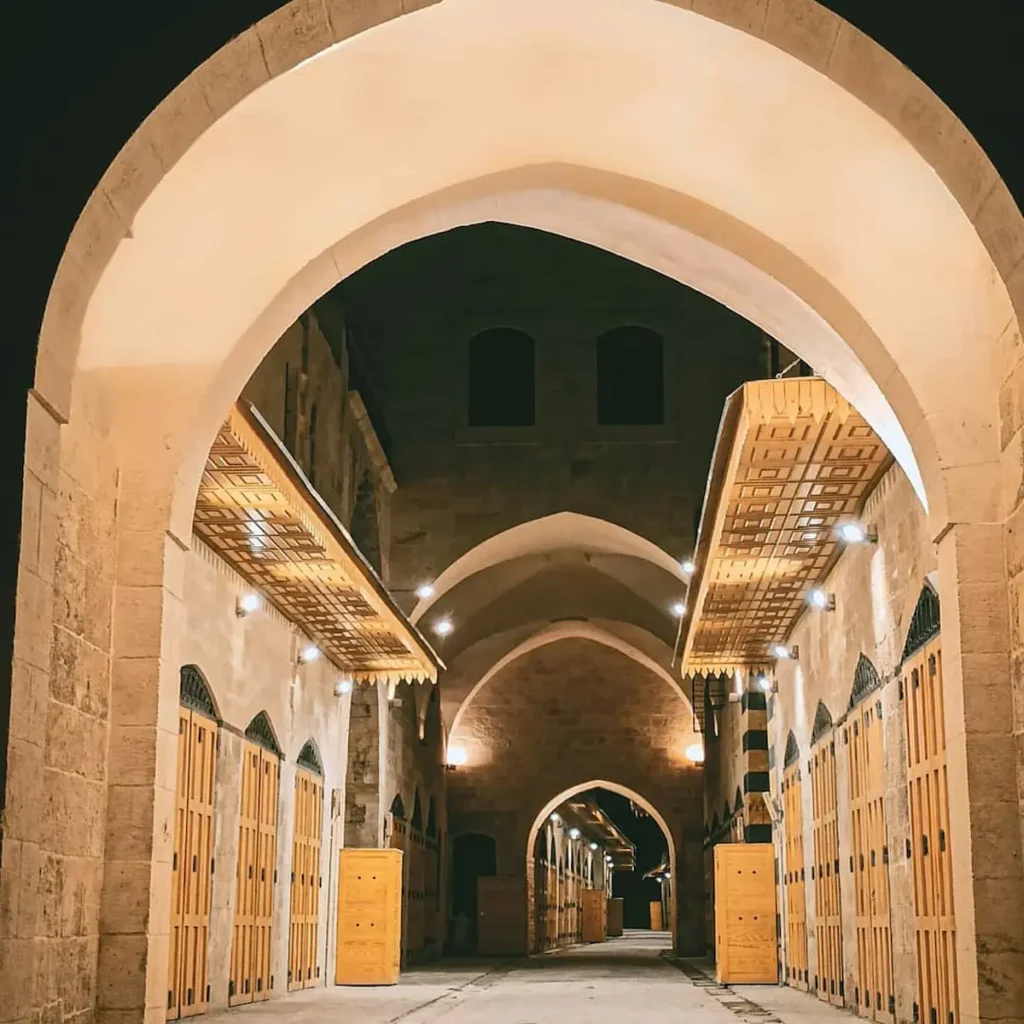 A nighttime view through a large stone archway into the restored Khan al-Harir in Aleppo, featuring warm lighting, vaulted ceilings, and uniform wooden shop doors along a clean stone-paved alleyway.