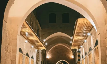 A nighttime view through a large stone archway into the restored Khan al-Harir in Aleppo, featuring warm lighting, vaulted ceilings, and uniform wooden shop doors along a clean stone-paved alleyway.