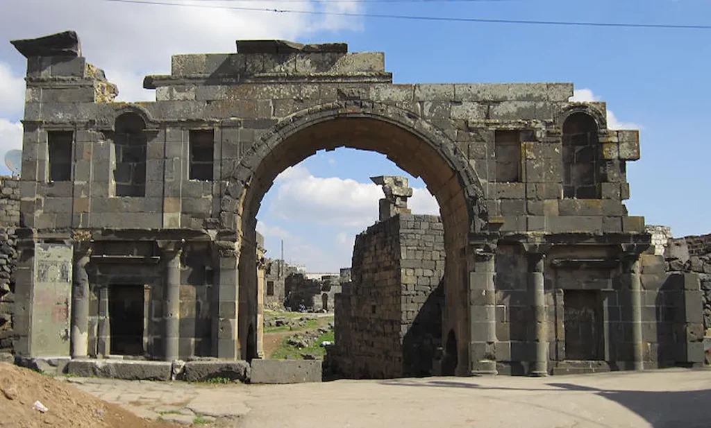 A large, ancient Roman triumphal arch made of dark black basalt stone, featuring a central high archway and smaller side niches, standing in the ruined city of Bosra under a blue sky.