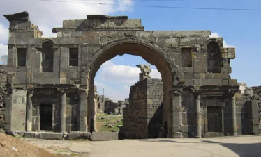 A large, ancient Roman triumphal arch made of dark black basalt stone, featuring a central high archway and smaller side niches, standing in the ruined city of Bosra under a blue sky.