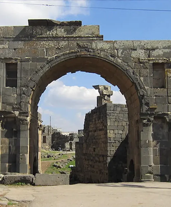 A large, ancient Roman triumphal arch made of dark black basalt stone, featuring a central high archway and smaller side niches, standing in the ruined city of Bosra under a blue sky.