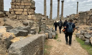 A group of travelers walking along a dirt path through the black basalt ruins of the Old City of Bosra, with towering ancient columns standing against an overcast sky in the background.