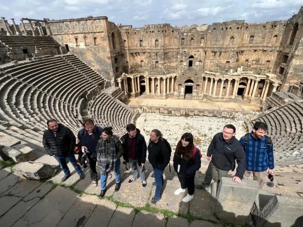 A high-angle group photo of eight travelers standing on the ancient stone steps of the Roman Theatre of Bosra, with the massive semi-circular seating and the ornate stone stage backdrop visible in the background.