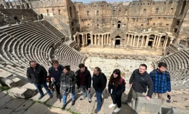 A high-angle group photo of eight travelers standing on the ancient stone steps of the Roman Theatre of Bosra, with the massive semi-circular seating and the ornate stone stage backdrop visible in the background.