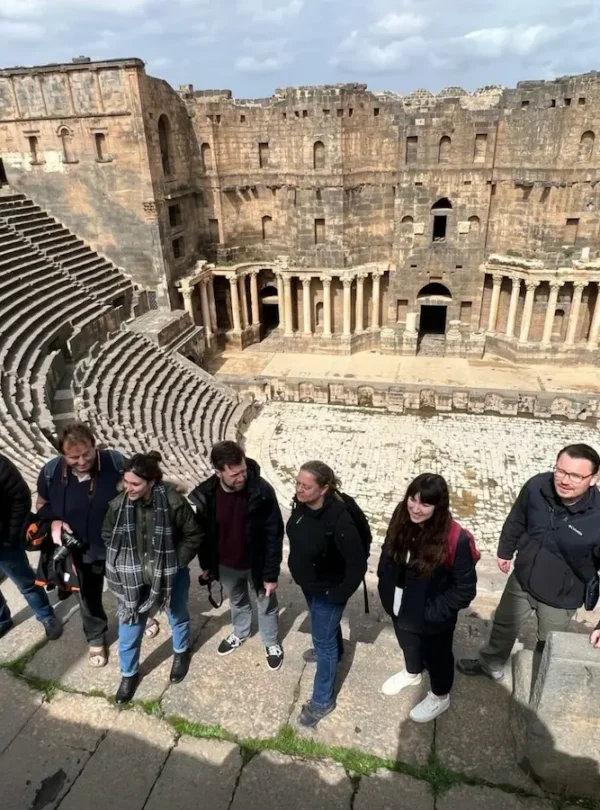 A high-angle group photo of eight travelers standing on the ancient stone steps of the Roman Theatre of Bosra, with the massive semi-circular seating and the ornate stone stage backdrop visible in the background.