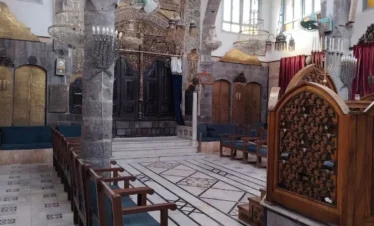 The interior of a historic synagogue in Damascus featuring high stone arches, ornate wooden screens with geometric carvings, rows of blue cushioned chairs, and multiple large, intricate glass chandeliers hanging from a beamed ceiling.