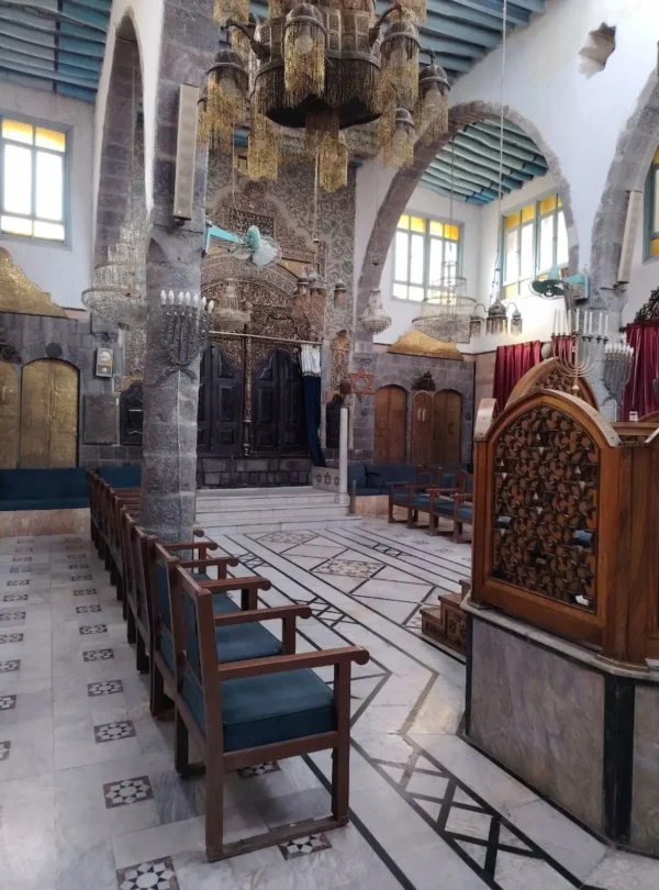 The interior of a historic synagogue in Damascus featuring high stone arches, ornate wooden screens with geometric carvings, rows of blue cushioned chairs, and multiple large, intricate glass chandeliers hanging from a beamed ceiling.