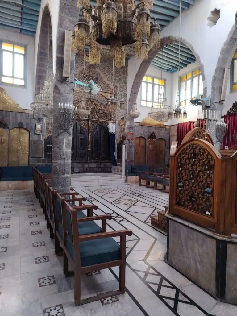The interior of a historic synagogue in Damascus featuring high stone arches, ornate wooden screens with geometric carvings, rows of blue cushioned chairs, and multiple large, intricate glass chandeliers hanging from a beamed ceiling.