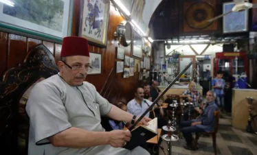 A man wearing a traditional red tarbush and grey robe sits in a wood-paneled café, holding a sword and reading from a small book to a group of men sitting in the background with hookahs.