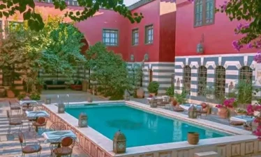 A high-angle view of a luxurious courtyard featuring a large turquoise swimming pool surrounded by a patterned stone patio with café tables, vibrant pink bougainvillea, and a grand red and white striped historic building.