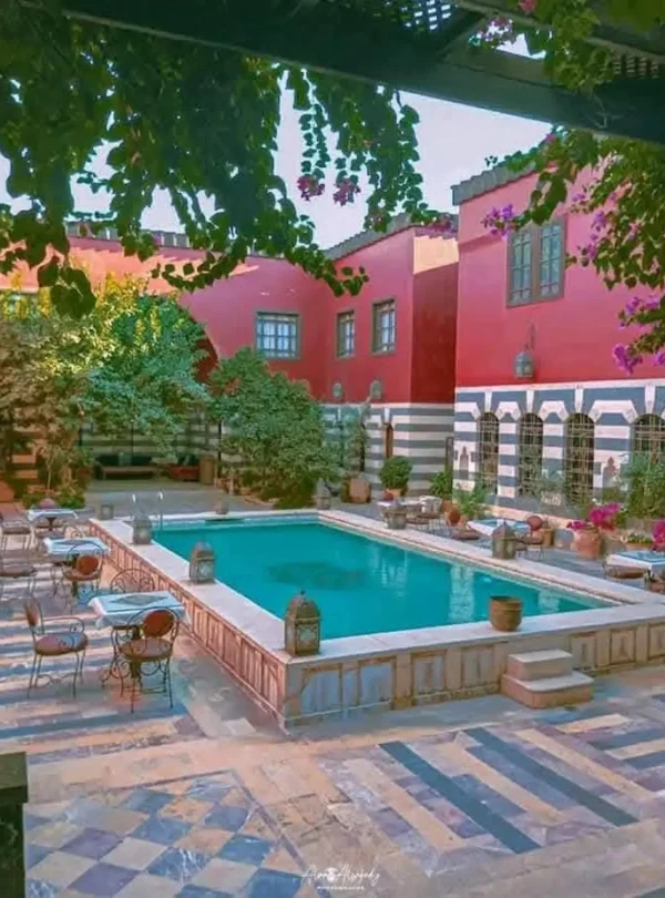 A high-angle view of a luxurious courtyard featuring a large turquoise swimming pool surrounded by a patterned stone patio with café tables, vibrant pink bougainvillea, and a grand red and white striped historic building.