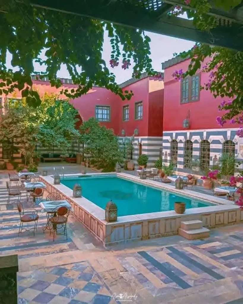 A high-angle view of a luxurious courtyard featuring a large turquoise swimming pool surrounded by a patterned stone patio with café tables, vibrant pink bougainvillea, and a grand red and white striped historic building.