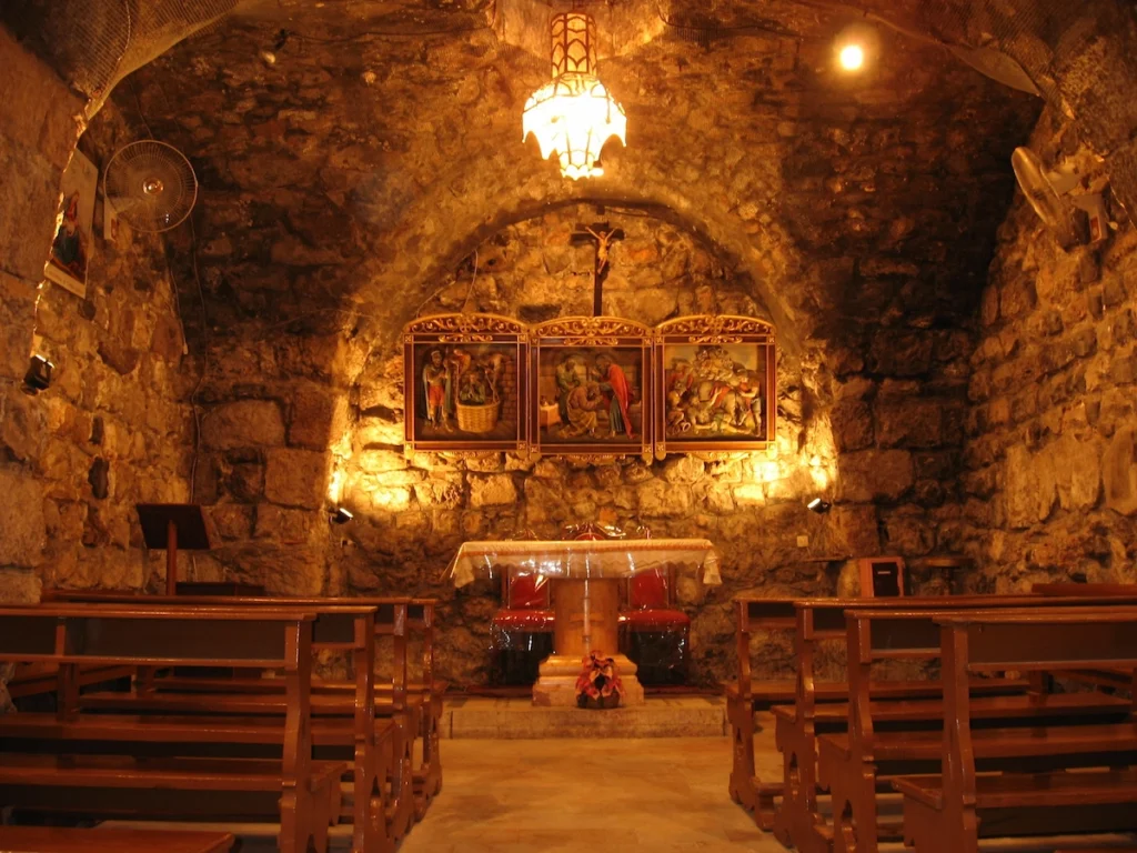 The interior of an ancient underground stone chapel with a low vaulted ceiling, featuring wooden pews, a simple altar, and illuminated religious icons on the far wall under a warm hanging light.