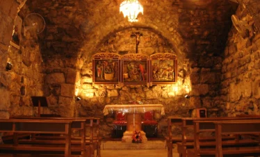 The interior of an ancient underground stone chapel with a low vaulted ceiling, featuring wooden pews, a simple altar, and illuminated religious icons on the far wall under a warm hanging light.
