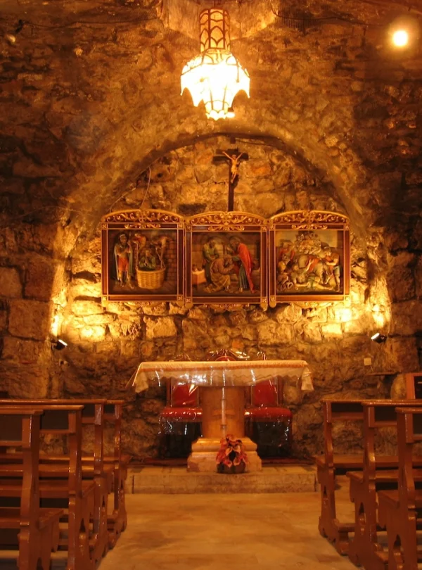 The interior of an ancient underground stone chapel with a low vaulted ceiling, featuring wooden pews, a simple altar, and illuminated religious icons on the far wall under a warm hanging light.