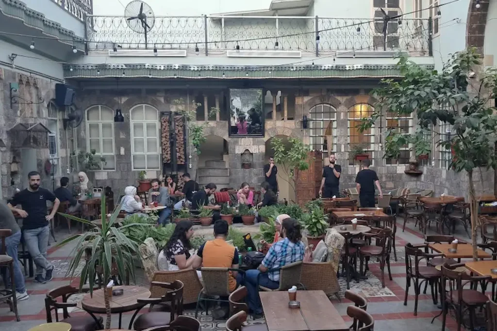 A bustling outdoor courtyard of a traditional stone building converted into a café, with people sitting at wooden tables among lush potted plants and string lights under a twilight sky.
