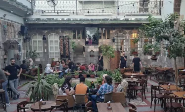 A bustling outdoor courtyard of a traditional stone building converted into a café, with people sitting at wooden tables among lush potted plants and string lights under a twilight sky.