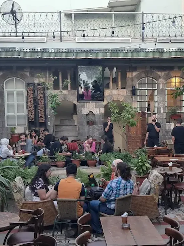 A bustling outdoor courtyard of a traditional stone building converted into a café, with people sitting at wooden tables among lush potted plants and string lights under a twilight sky.