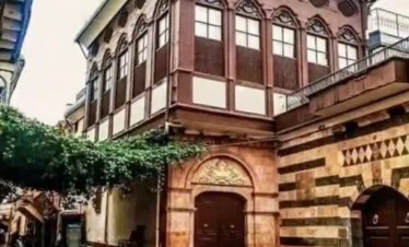 A narrow, wet cobblestone street in Old Damascus featuring a striking two-story building with a dark wooden upper floor and ornate pointed windows, adjacent to a stone wall with traditional red and white horizontal stripes.