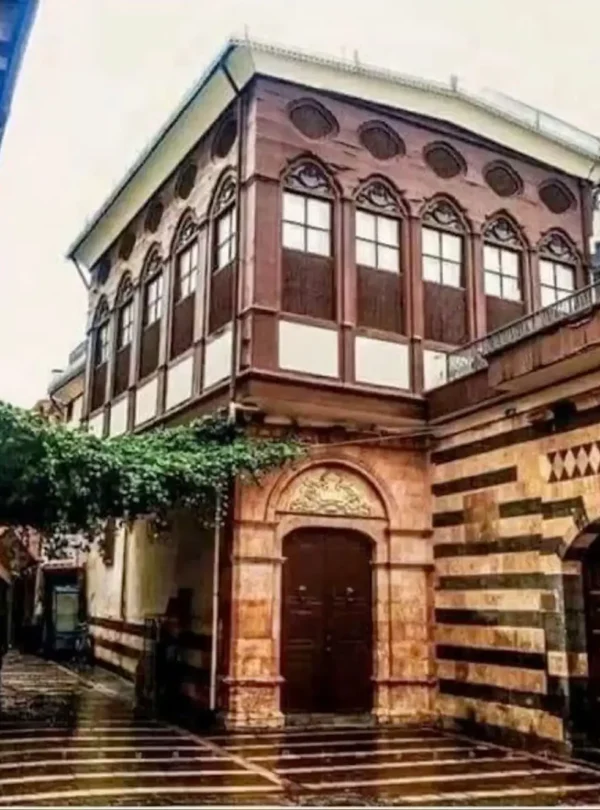 A narrow, wet cobblestone street in Old Damascus featuring a striking two-story building with a dark wooden upper floor and ornate pointed windows, adjacent to a stone wall with traditional red and white horizontal stripes.