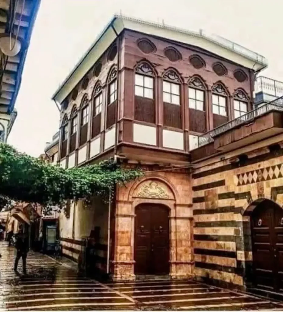 A narrow, wet cobblestone street in Old Damascus featuring a striking two-story building with a dark wooden upper floor and ornate pointed windows, adjacent to a stone wall with traditional red and white horizontal stripes.