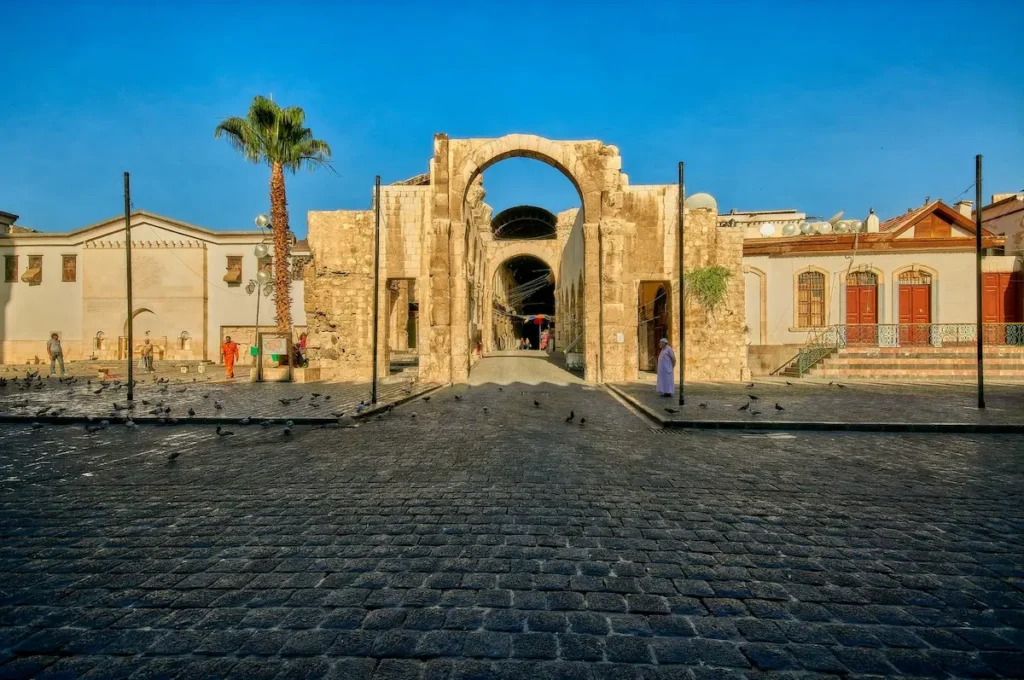 A wide view of an ancient Roman stone triumphal arch standing in a large cobblestone plaza in Old Damascus, under a clear blue sky with a tall palm tree to the left.