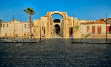 A wide view of an ancient Roman stone triumphal arch standing in a large cobblestone plaza in Old Damascus, under a clear blue sky with a tall palm tree to the left.