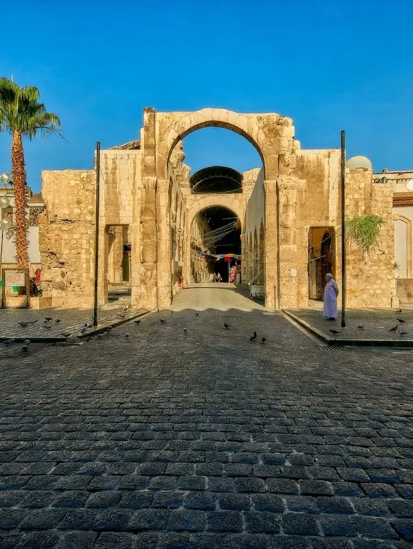 A wide view of an ancient Roman stone triumphal arch standing in a large cobblestone plaza in Old Damascus, under a clear blue sky with a tall palm tree to the left.