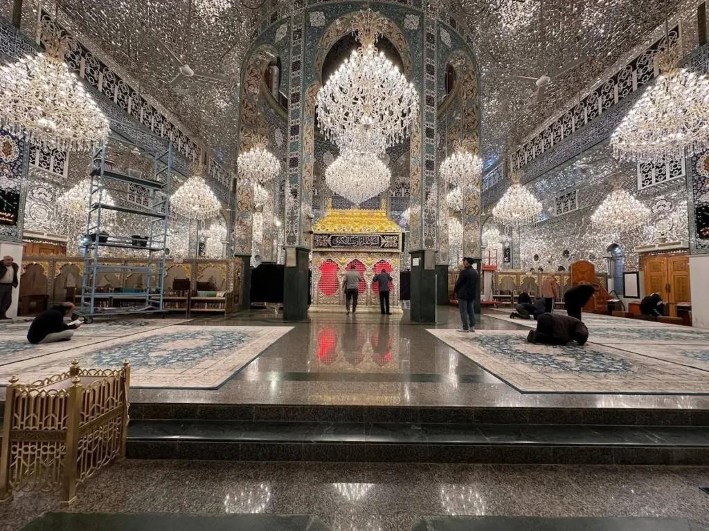 The interior of a grand shrine in Damascus, featuring walls and ceilings covered in intricate silver mirror mosaics, large crystal chandeliers, and a central gold-roofed tomb surrounded by blue patterned carpets.