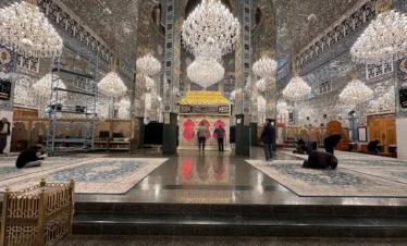 The interior of a grand shrine in Damascus, featuring walls and ceilings covered in intricate silver mirror mosaics, large crystal chandeliers, and a central gold-roofed tomb surrounded by blue patterned carpets.