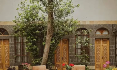 A tranquil interior courtyard of a traditional Damascene house featuring a central marble fountain, vibrant orange and pink flowers in terracotta pots, and intricate wooden doors under a towering green tree.