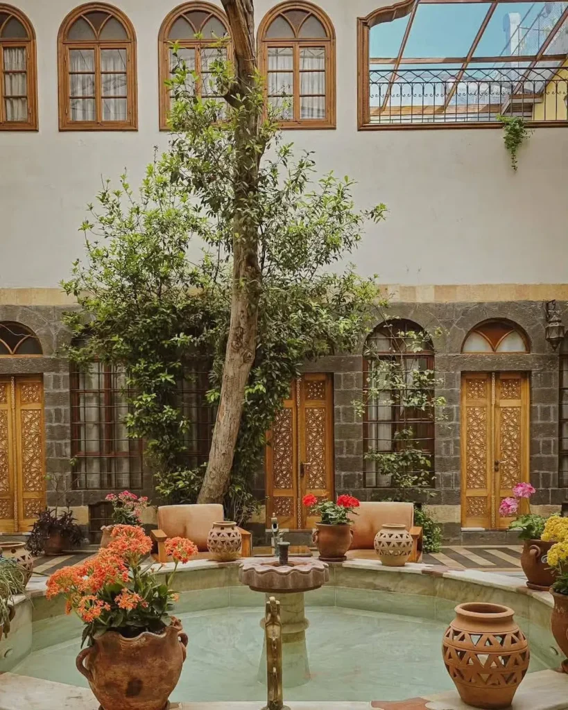 A tranquil interior courtyard of a traditional Damascene house featuring a central marble fountain, vibrant orange and pink flowers in terracotta pots, and intricate wooden doors under a towering green tree.