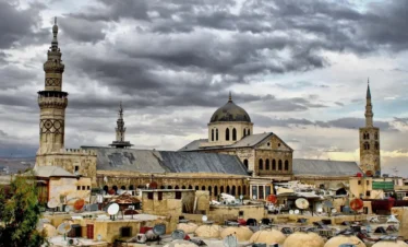 A high-angle view of the Umayyad Mosque in Damascus under a dramatic cloudy sky, showing its large central dome and distinctive minarets rising above the crowded rooftops of the Old City.