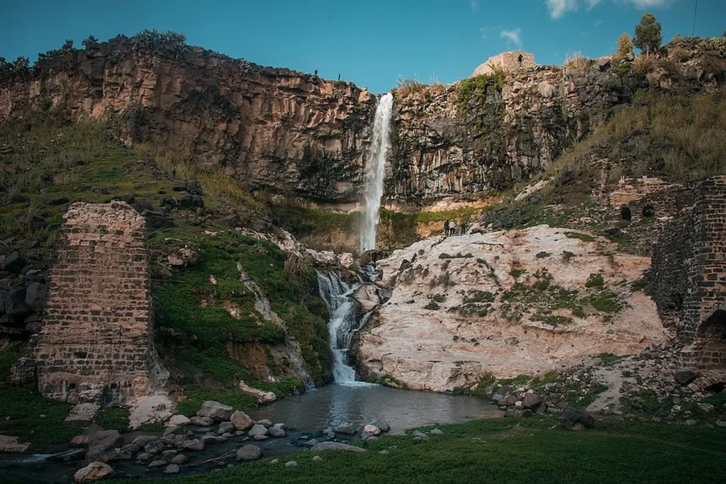 A tall, slender waterfall cascading down a steep rocky cliff into a small pool, surrounded by green hills and the ancient stone remains of a watermill in Tell Shihab, Syria.