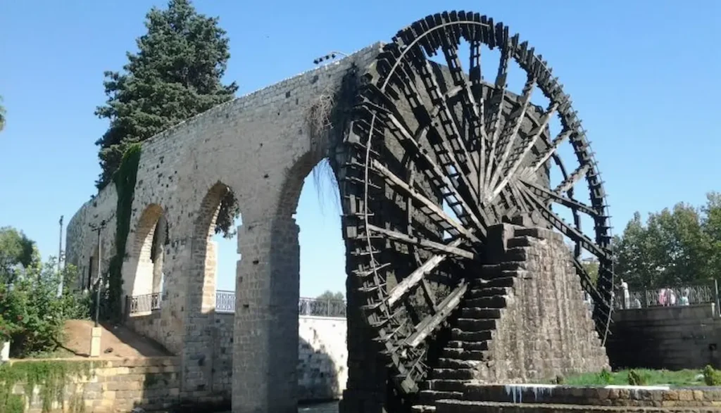 A massive, intricate wooden waterwheel (noria) attached to a high stone arched aqueduct over a river in Hama, Syria, under a clear blue sky.
