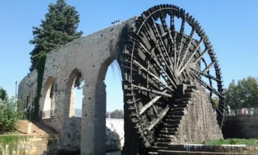 A massive, intricate wooden waterwheel (noria) attached to a high stone arched aqueduct over a river in Hama, Syria, under a clear blue sky.