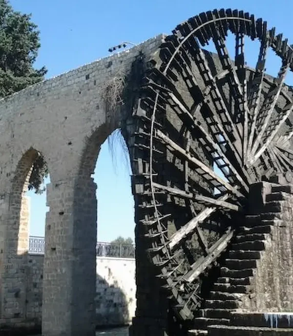 A massive, intricate wooden waterwheel (noria) attached to a high stone arched aqueduct over a river in Hama, Syria, under a clear blue sky.