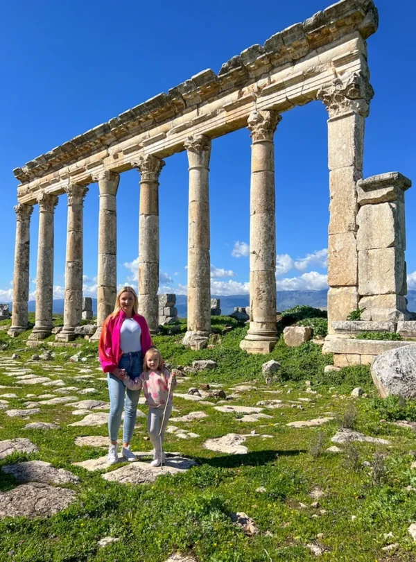 A woman in a pink jacket and a young child stand hand-in-hand on the ancient, stone-paved Cardo Maximus of Apamea, framed by a row of towering Roman columns under a bright blue sky.