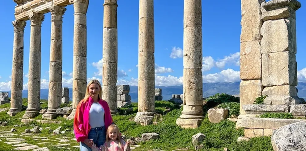 A woman in a pink jacket and a young child stand hand-in-hand on the ancient, stone-paved Cardo Maximus of Apamea, framed by a row of towering Roman columns under a bright blue sky.