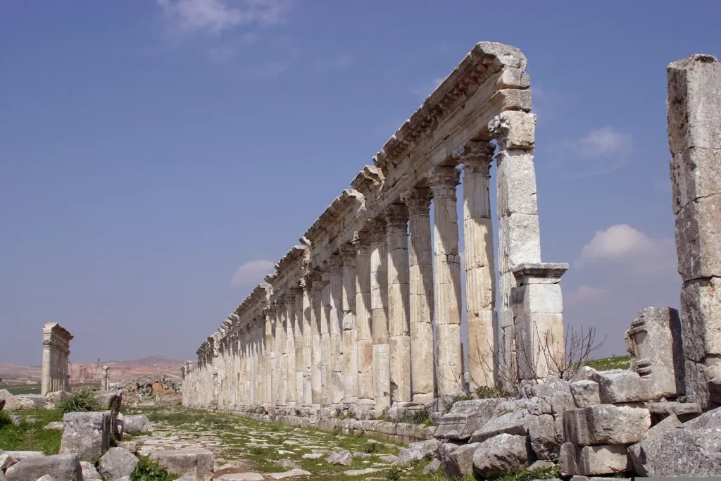 A low-angle perspective looking down an exceptionally long row of ancient fluted Roman columns with ornate Corinthian capitals, stretching toward the horizon under a bright blue sky with light clouds.
