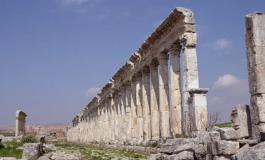 A low-angle perspective looking down an exceptionally long row of ancient fluted Roman columns with ornate Corinthian capitals, stretching toward the horizon under a bright blue sky with light clouds.