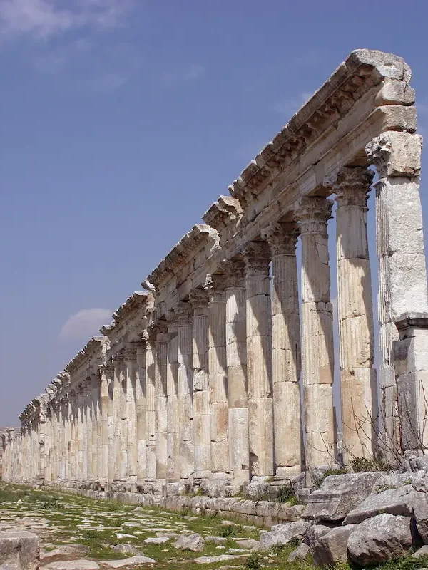 A low-angle perspective looking down an exceptionally long row of ancient fluted Roman columns with ornate Corinthian capitals, stretching toward the horizon under a bright blue sky with light clouds.
