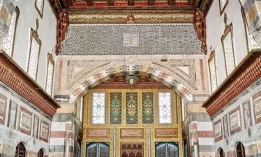 The highly ornate interior of a grand Ottoman palace room featuring striped stone walls, an intricately carved and painted wooden ceiling, geometric tile patterns, and low cushioned seating along the walls.