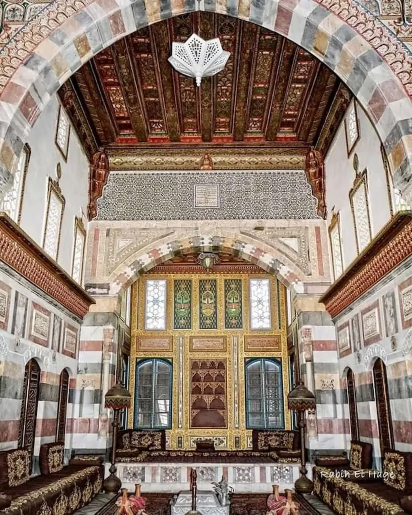 The highly ornate interior of a grand Ottoman palace room featuring striped stone walls, an intricately carved and painted wooden ceiling, geometric tile patterns, and low cushioned seating along the walls.