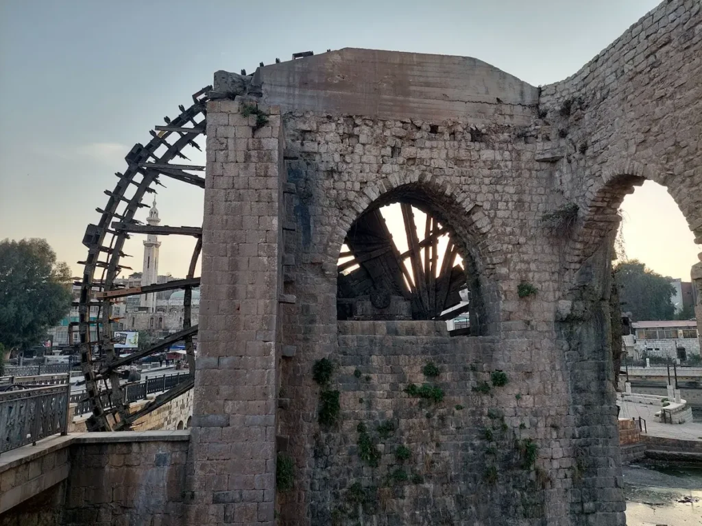 A side view of a massive wooden waterwheel (noria) attached to a medieval stone structure in Hama, with the minaret of a mosque visible in the background against a pale dusk sky.
