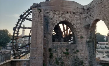 A side view of a massive wooden waterwheel (noria) attached to a medieval stone structure in Hama, with the minaret of a mosque visible in the background against a pale dusk sky.