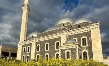 A large mosque with dark basalt stone walls, silver domes, and a tall, slender minaret, viewed from behind a field of bright yellow wildflowers under a dramatic, cloudy blue sky.