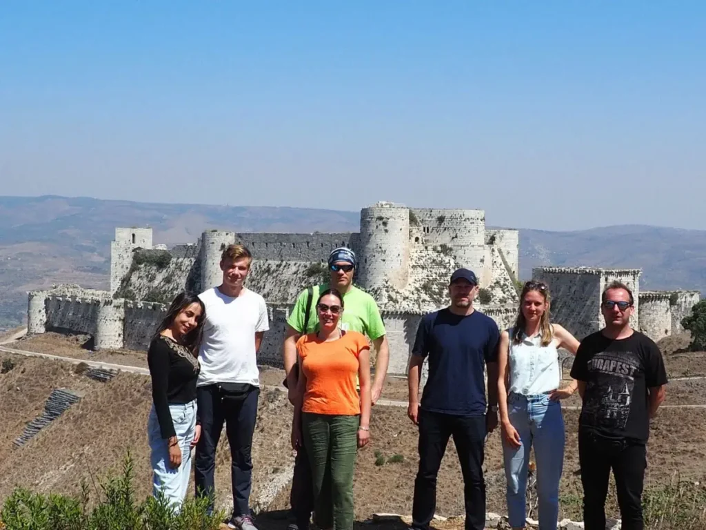 A group of six travelers in casual summer clothing stands on a hillside, posing in front of the massive stone walls and round towers of the Krak des Chevaliers crusader castle under a clear blue sky.